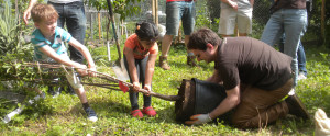 Two children pull a small tree from a container that a man is holding.